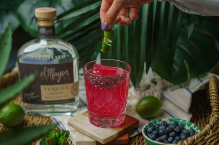 Hand garnishing a vibrant blueberry margarita with lime peel, alongside El Mayor tequila, limes, and blueberries.