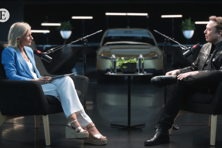 Elon Musk and a woman in blue blazer seated with microphones, golden car in background