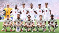 England football team players celebrate on the field with fans in the background during an international match.