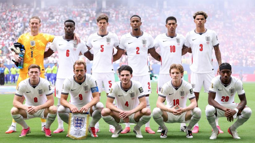 England football team players celebrate on the field with fans in the background during an international match.