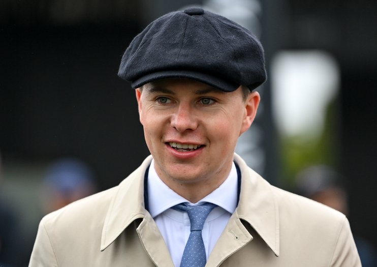 Smiling man in flat cap, light trench coat, white shirt, and blue tie, looking right.