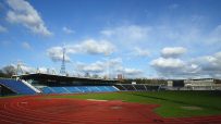 Outdoor athletic stadium with red running track, green field, blue spectator seating, and large scoreboards.