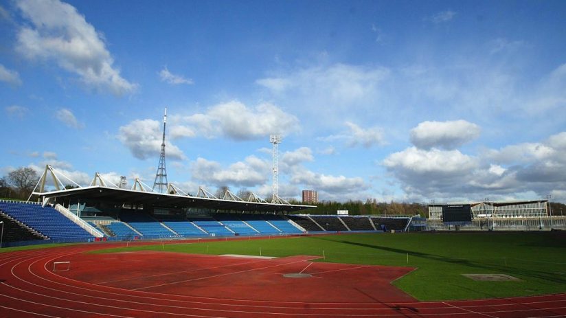 Outdoor athletic stadium with red running track, green field, blue spectator seating, and large scoreboards.