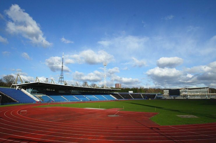 Outdoor athletic stadium with red running track, green field, blue spectator seating, and large scoreboards.