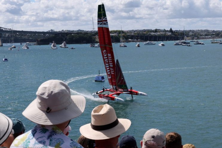 Red foiling sailboat racing on blue water with a bridge and city in the background, spectators watching