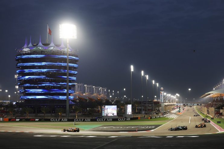 Night race at Bahrain International Circuit with brightly lit tower, grandstands, and F1 cars on track.
