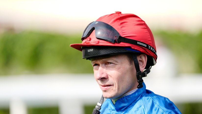 Jockey Ryan Moore in a red helmet and blue silks, wearing sunglasses, looking focused at a racecourse