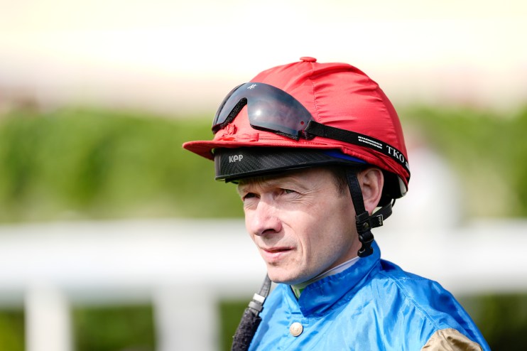 Jockey Ryan Moore in a red helmet and blue silks, wearing sunglasses, looking focused at a racecourse