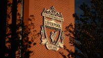 Liverpool FC crest on a red brick wall, illuminated by sunlight with tree shadows.