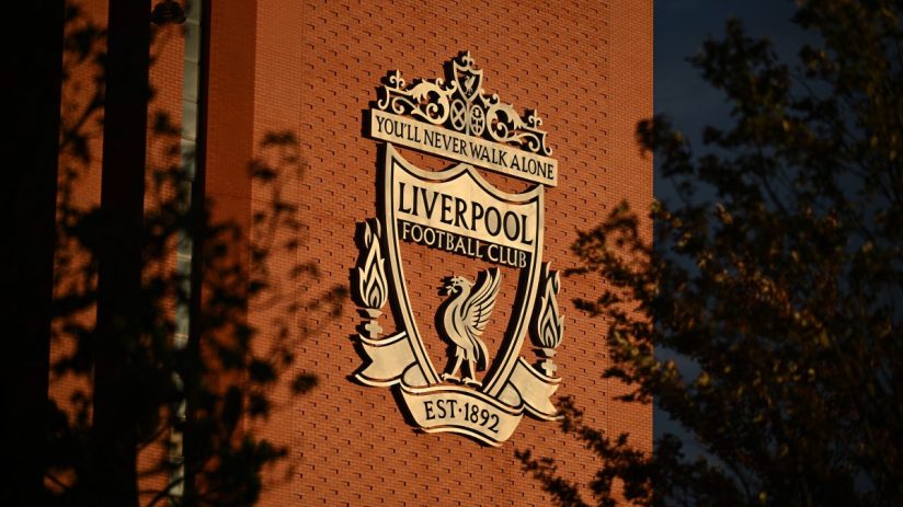 Liverpool FC crest on a red brick wall, illuminated by sunlight with tree shadows.