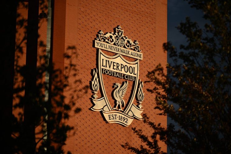 Liverpool FC crest on a red brick wall, illuminated by sunlight with tree shadows.