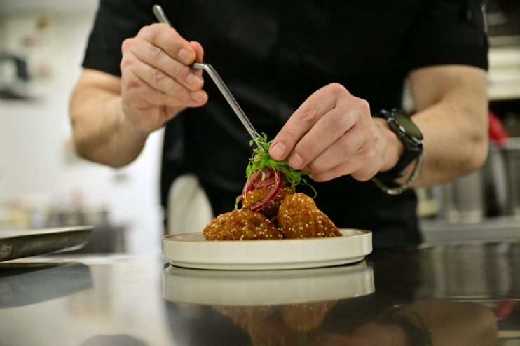 Chef garnishing falafel with microgreens and pickled onions using tweezers on a white plate in a kitchen