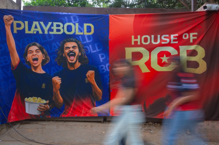 Blue and red banners featuring Playbold and House of RCB with cheering fans, blurred figures passing by.