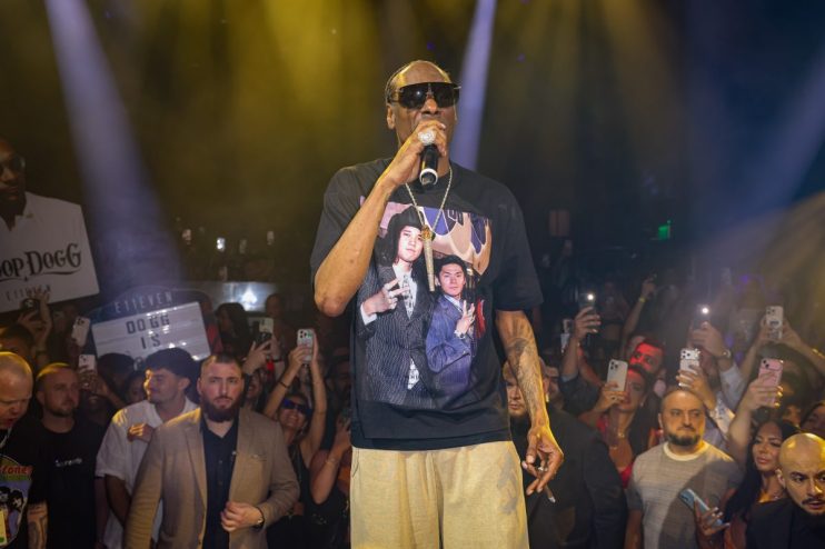 Snoop Dogg performing on stage, holding a microphone, wearing sunglasses and a graphic t-shirt, surrounded by a crowd