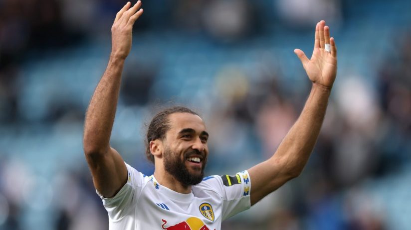 Happy male soccer player with beard and long hair in white uniform raising arms, smiling