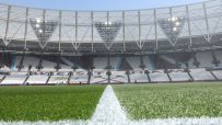 Football pitch with a white line, stadium seats, and bright lights under a clear sky.