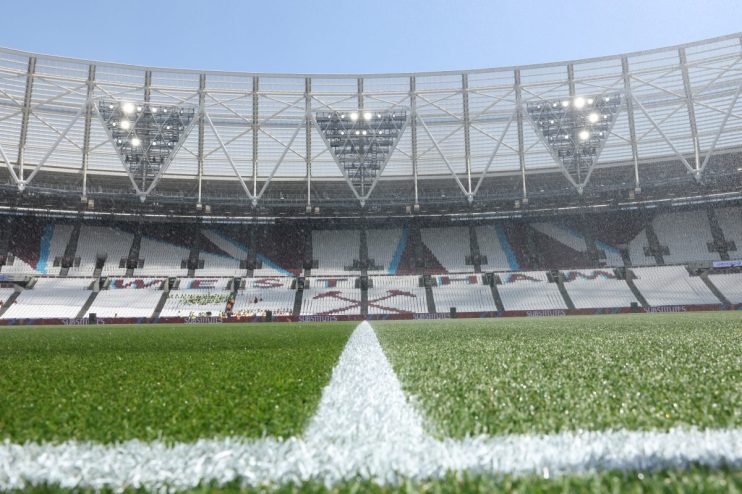 Football pitch with a white line, stadium seats, and bright lights under a clear sky.