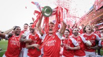 Arsenal players celebrate with the Premier League trophy and confetti raining down, cheering on the field.