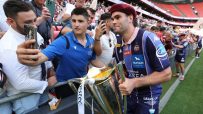 Rugby player in a maroon beret and blue jersey holding a trophy, surrounded by fans taking selfies at a stadium.