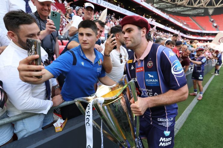 Rugby player in a maroon beret and blue jersey holding a trophy, surrounded by fans taking selfies at a stadium.