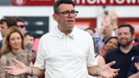 Andy Burnham speaking at a podium with microphones, gesturing with his hands, crowd in background