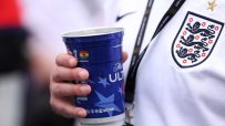 Hand holding a blue FIFA World Cup 2026 sponsored cup, with an England football jersey visible.