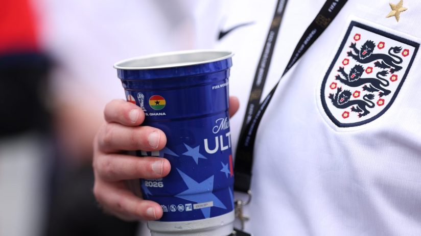 Hand holding a blue FIFA World Cup 2026 sponsored cup, with an England football jersey visible.