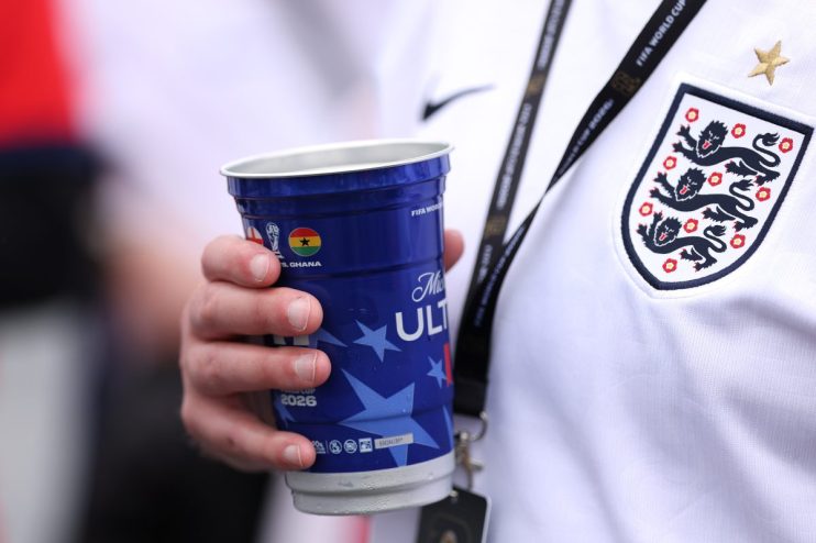 Hand holding a blue FIFA World Cup 2026 sponsored cup, with an England football jersey visible.