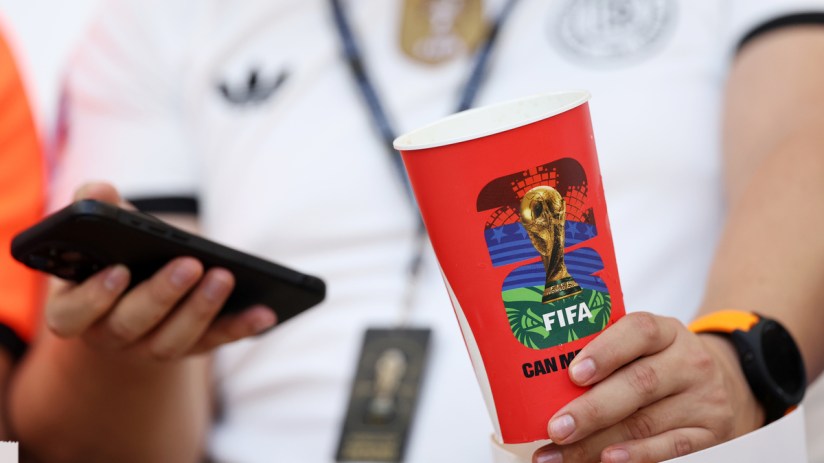 Person holding a red FIFA World Cup branded cup and a smartphone, wearing a white shirt and watch
