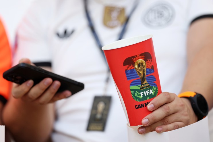 Person holding a red FIFA World Cup branded cup and a smartphone, wearing a white shirt and watch