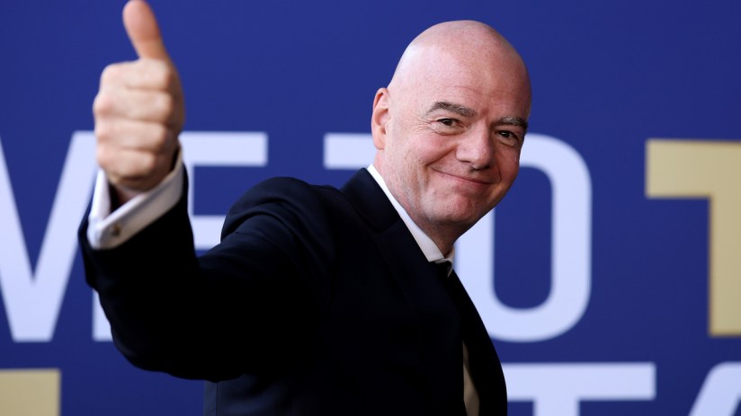 Gianni Infantino smiling and giving a thumbs up, wearing a suit against a blue and white background.