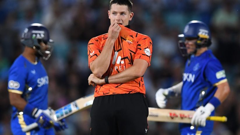 A male cricketer in an orange and black uniform looks concerned during a match, surrounded by other players.