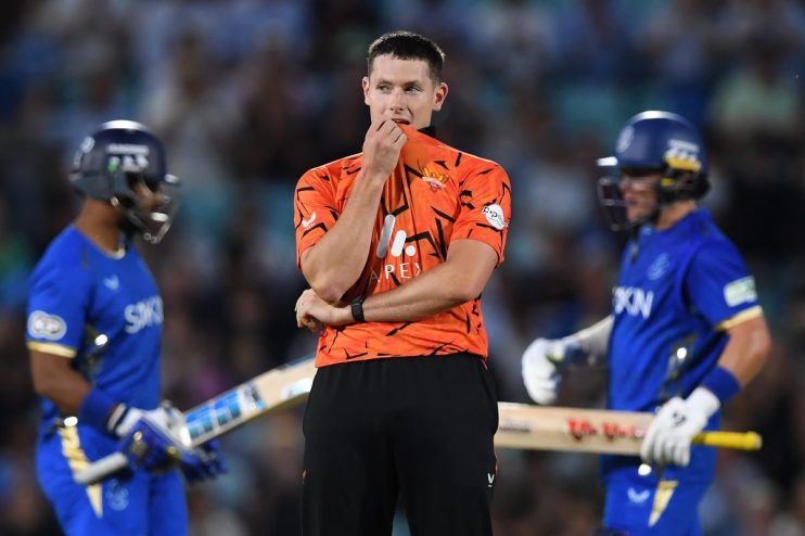 A male cricketer in an orange and black uniform looks concerned during a match, surrounded by other players.