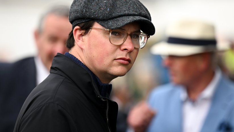 A man in a tweed flat cap and glasses looks toward the viewer, with blurred figures in the background.