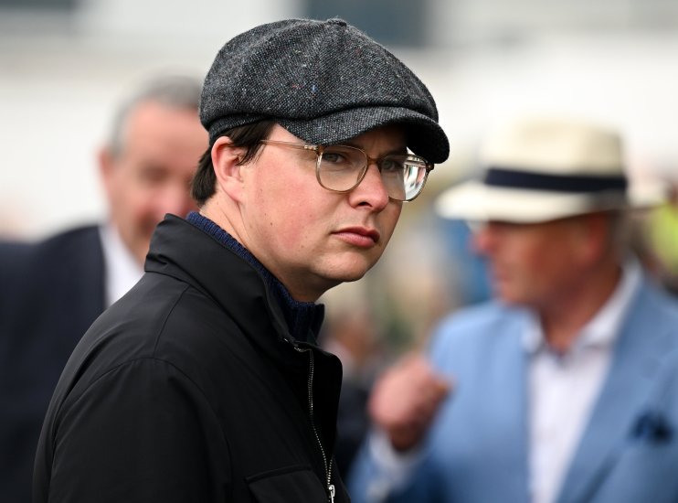 A man in a tweed flat cap and glasses looks toward the viewer, with blurred figures in the background.