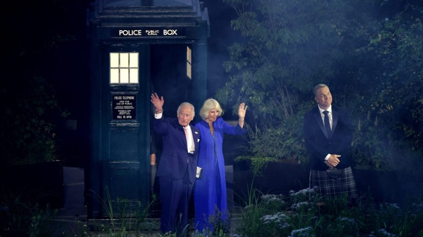 King Charles III and Queen Camilla wave from a TARDIS police box, with a man in a kilt nearby.