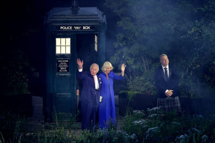King Charles III and Queen Camilla wave from a TARDIS police box, with a man in a kilt nearby.