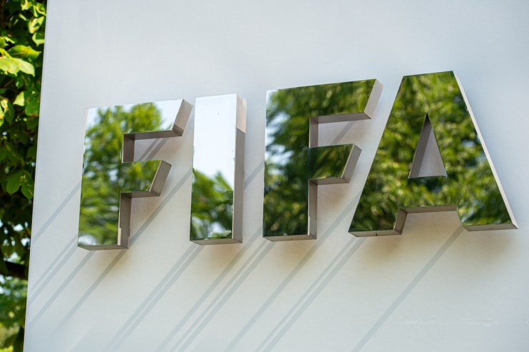 Reflective FIFA logo sign on a white wall, with green trees mirrored in the letters, under natural light.