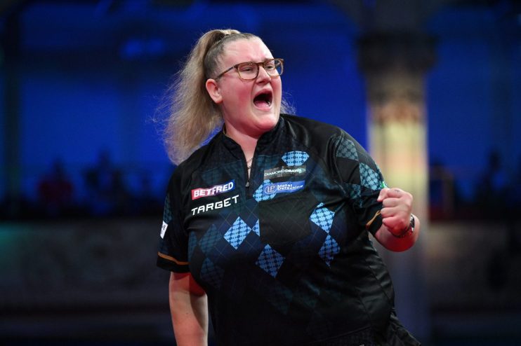 Blonde woman in glasses and a patterned shirt, with her mouth open and fist clenched, celebrating during a darts match