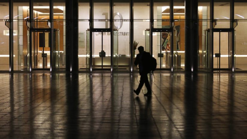 Silhouetted person walks past a modern building with 10 Upper Bank Street visible on its glass facade at night.