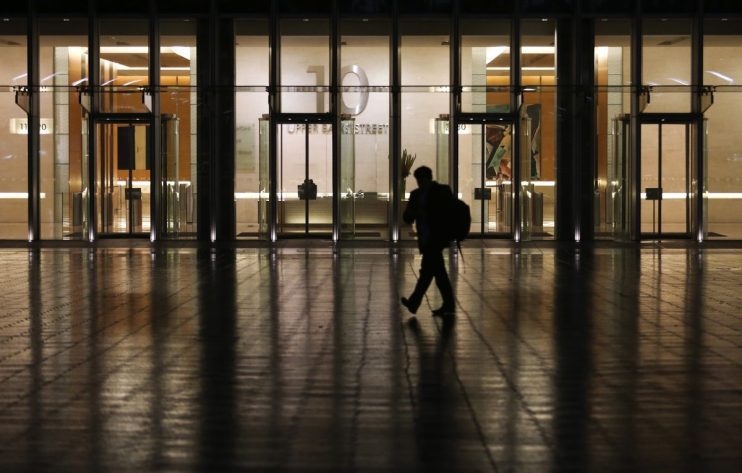 Silhouetted person walks past a modern building with 10 Upper Bank Street visible on its glass facade at night.
