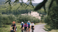 Cyclists ride on a winding road with a historic building and forested hills in Girona, Spain