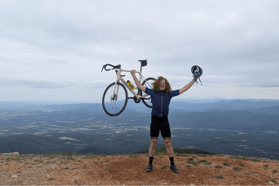 Excited cyclist holding up their bike and helmet on a mountain peak overlooking the Girona landscape.