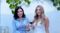 Two women smiling, one in blue holding white Greek wine, another in white holding red wine, under grapevines.