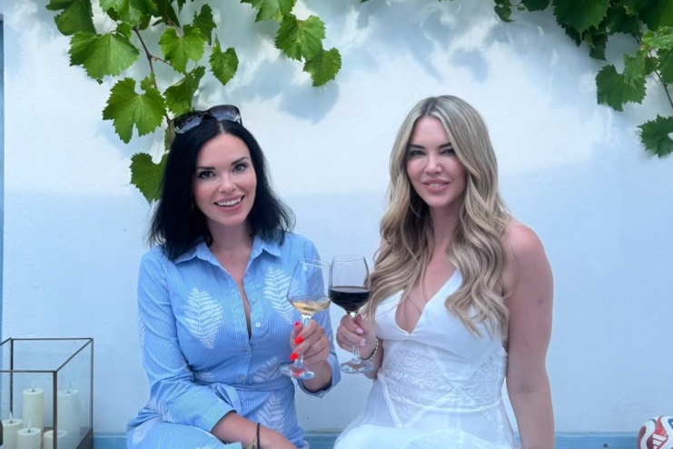 Two women smiling, one in blue holding white Greek wine, another in white holding red wine, under grapevines.