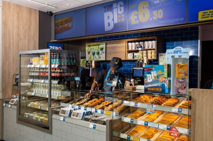 Interior of a Greggs bakery with a staff member behind the counter, displays of pastries, drinks, and The Big Deal signage.