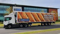 White Greggs delivery truck with Nations Favourite Sausage Roll graphic, parked outside a modern building.