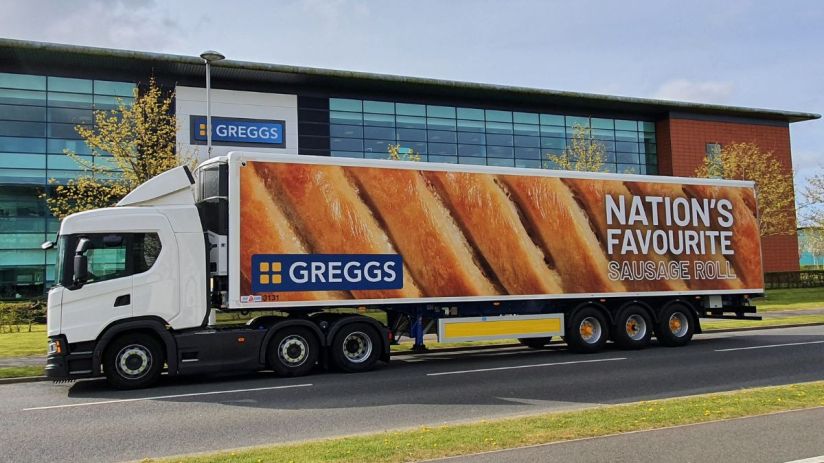 White Greggs delivery truck with Nations Favourite Sausage Roll graphic, parked outside a modern building.