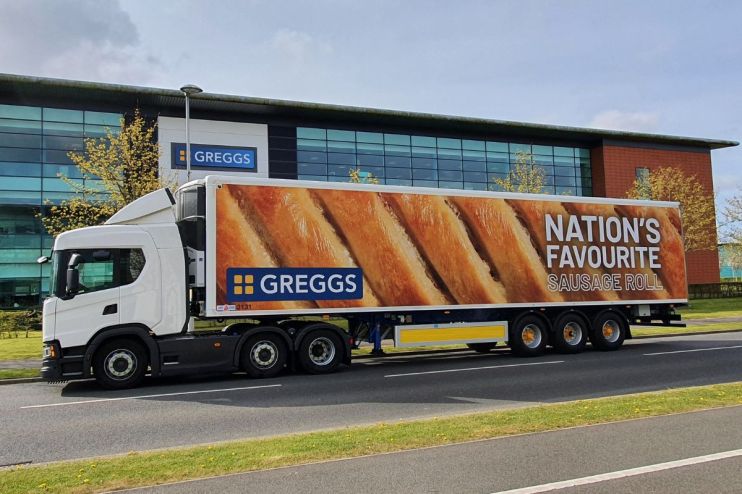 White Greggs delivery truck with Nations Favourite Sausage Roll graphic, parked outside a modern building.