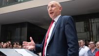 Man in suit and red tie speaking at a podium to an audience in a modern building.